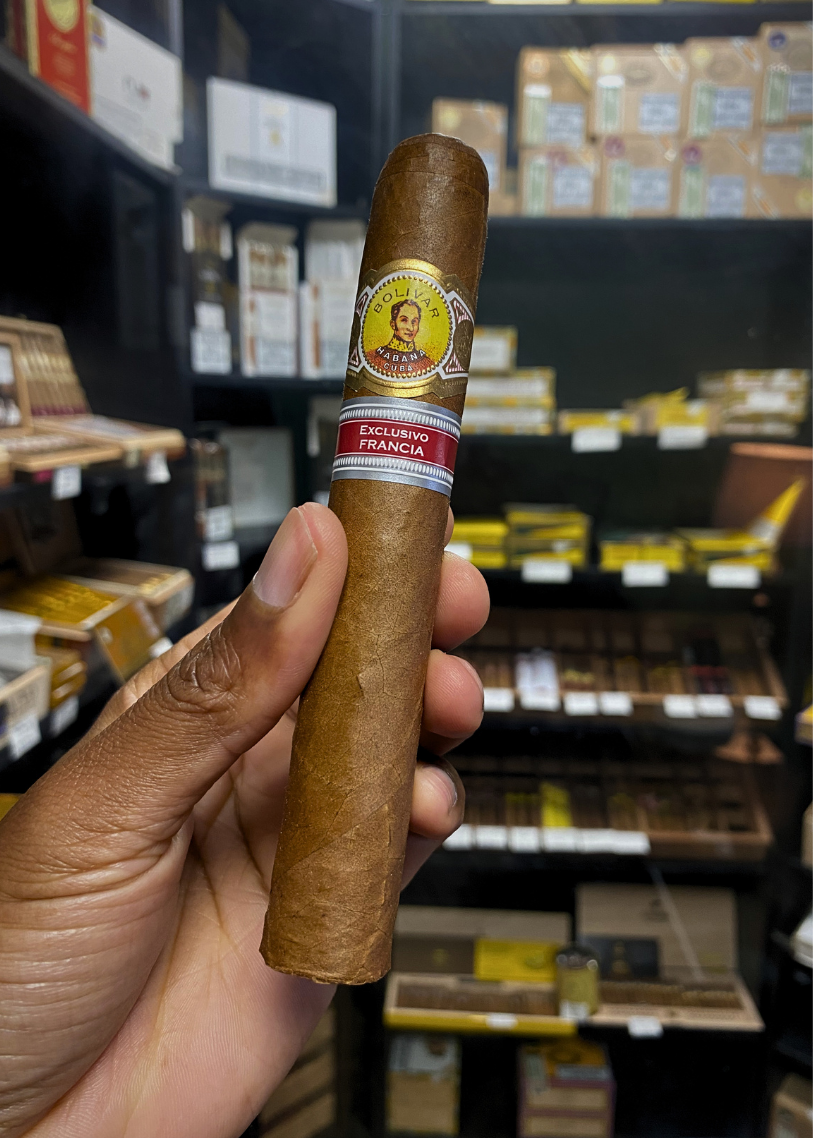 Cigar held by a hand with shelves of cigars in the background. Bolívar Libertador Regional Edition France 