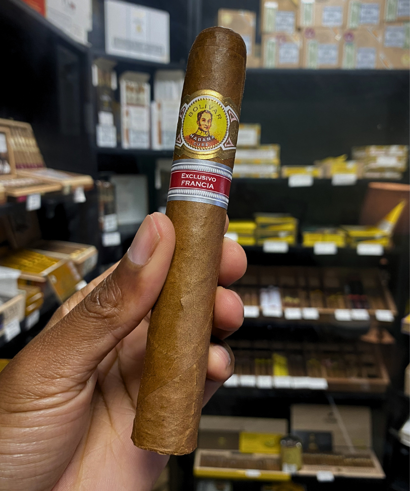 Cigar held by a hand with shelves of cigars in the background. Bolívar Libertador Regional Edition France 
