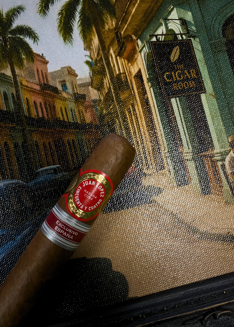 Cigar with a label held in front of a scenic painting of a street scene. Juan Lopez Cincuenta y Cuatro Regional Edition Spain 2025 