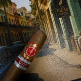 Cigar with a label held in front of a scenic painting of a street scene. Juan Lopez Cincuenta y Cuatro Regional Edition Spain 2025 