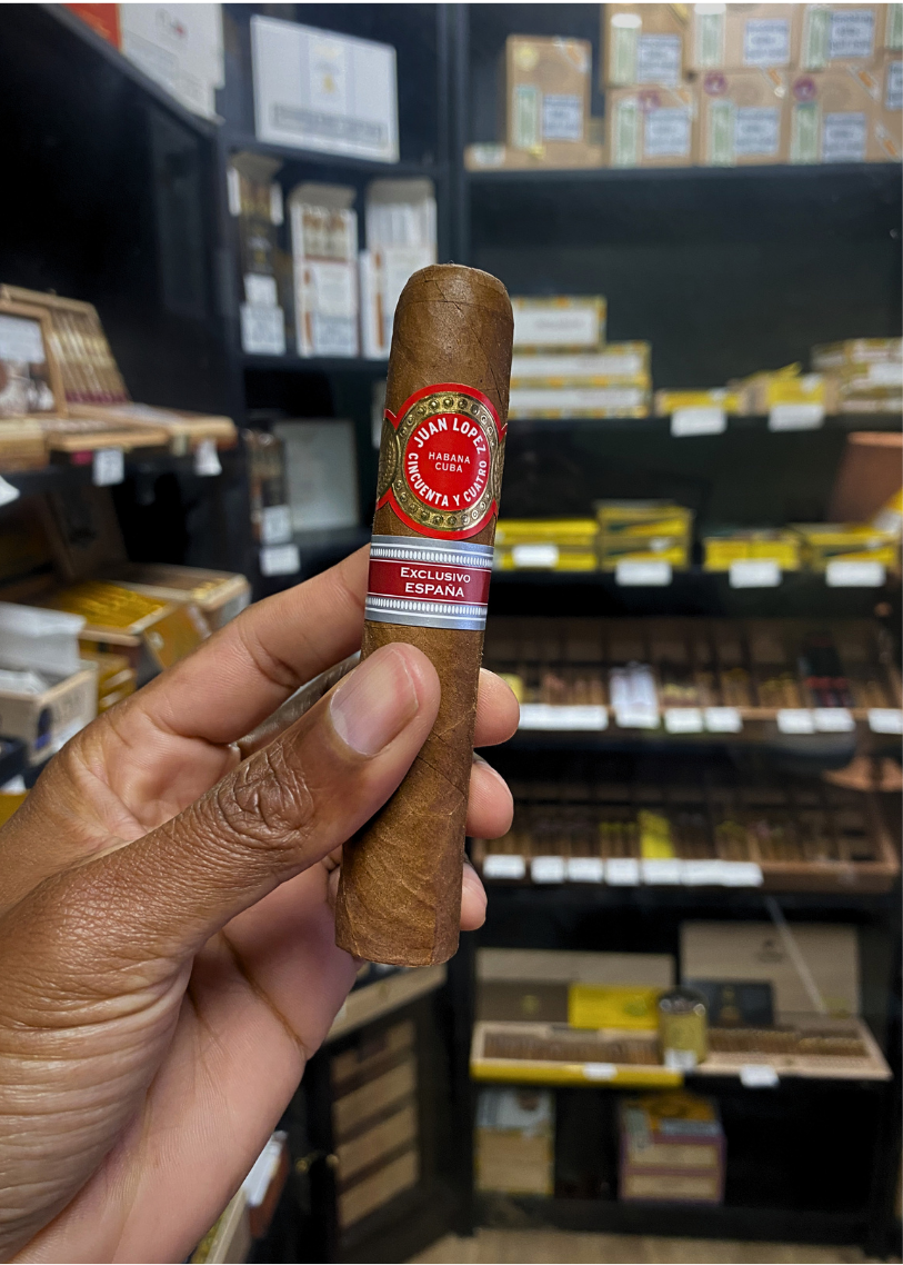 Cigar held by a hand with shelves of cigars in the background. Juan Lopez Cincuenta y Cuatro Regional Edition Spain 2025 