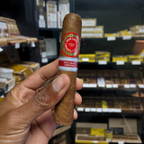 Cigar held by a hand with shelves of cigars in the background. Juan Lopez Cincuenta y Cuatro Regional Edition Spain 2025 