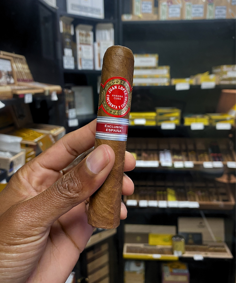 Cigar held by a hand with shelves of cigars in the background. Juan Lopez Cincuenta y Cuatro Regional Edition Spain 2025 