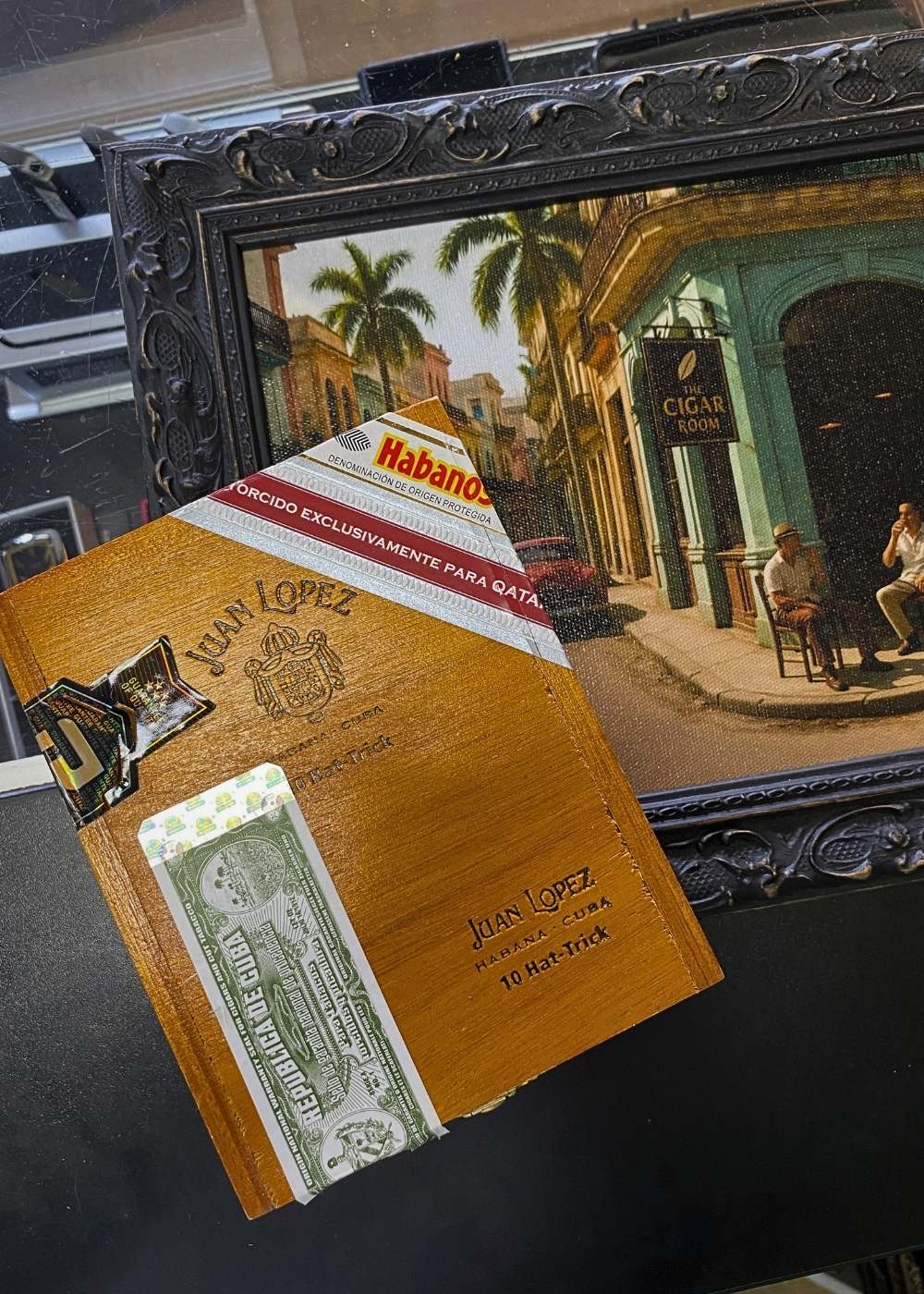Cigar box with Habano and Juan Lopez branding in front of a painting of a street scene.