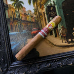 Cigar with a 'Vitello' label in front of a framed painting of a street scene.