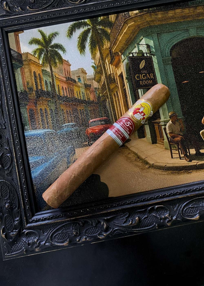 Cigar with a 'Vitello' label in front of a framed painting of a street scene.