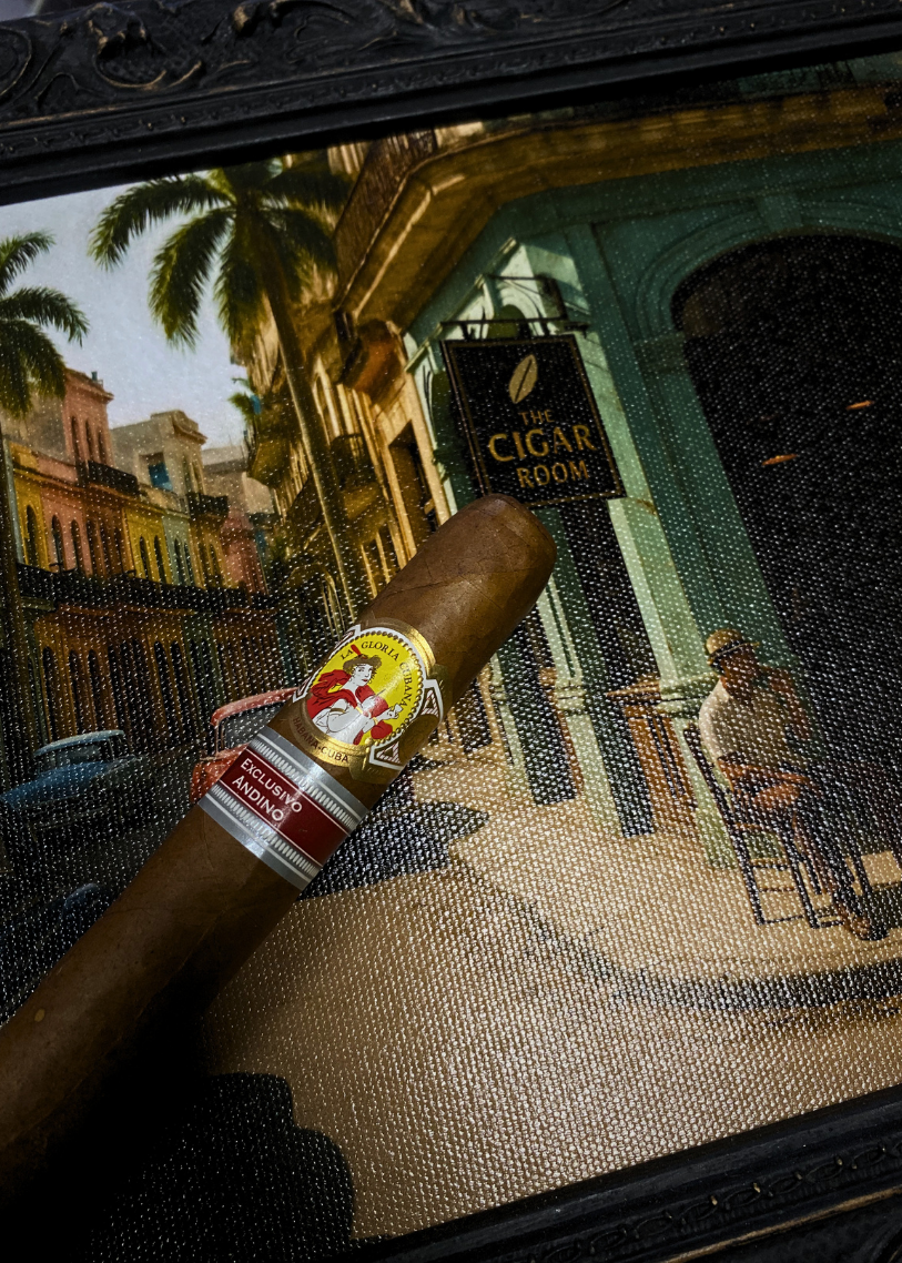 Cigar with band held over a painting of a street scene with 'The Cigar Room' sign.