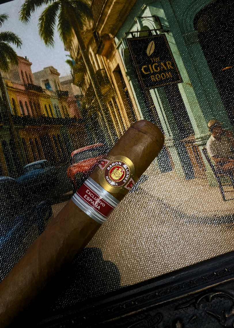 Cigar with a label held in front of a decorative background featuring a sign for 'The Cigar Room'. Ramón Allones Caprichos Regional Edition Spain