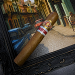 Cigar with a brand logo held in front of a scenic street view, possibly in a tropical or Latin American city.
