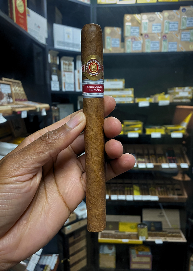 Cigar held in a hand with shelves of cigars in the background. Ramón Allones Grandes Regional Edition Spain