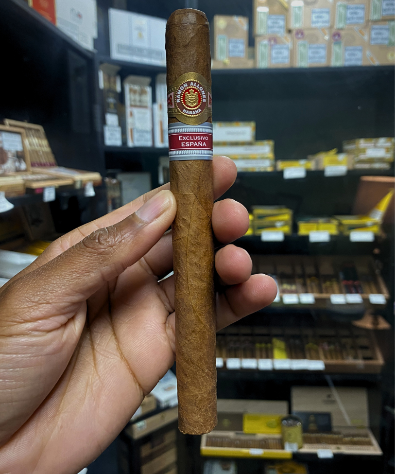 Cigar held in a hand with shelves of cigars in the background. Ramón Allones Grandes Regional Edition Spain