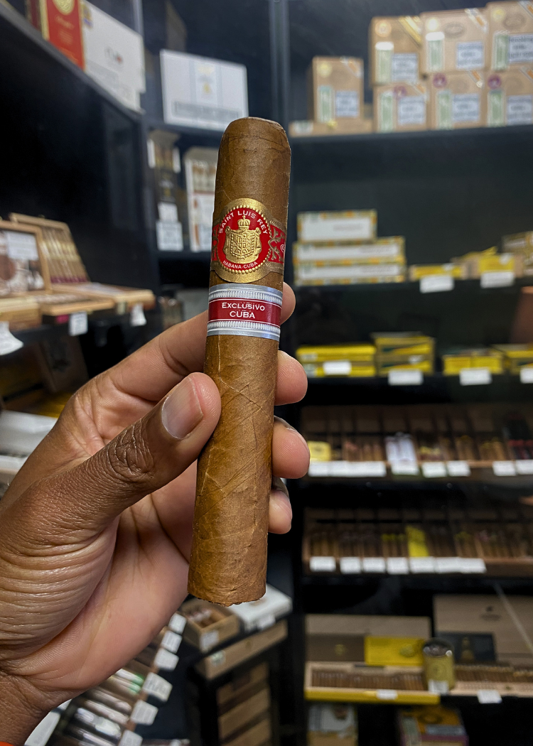 Cigar held by a hand with shelves of cigars in the background. Saint Luis Rey Marquéz Regional Edition Exclusive Cuba 2016