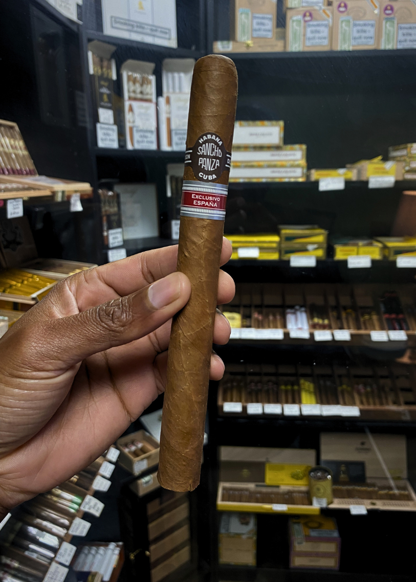 Cigar held by a hand with a store shelf of cigars in the background. Sancho Panza Quijote Regional Edition Spain