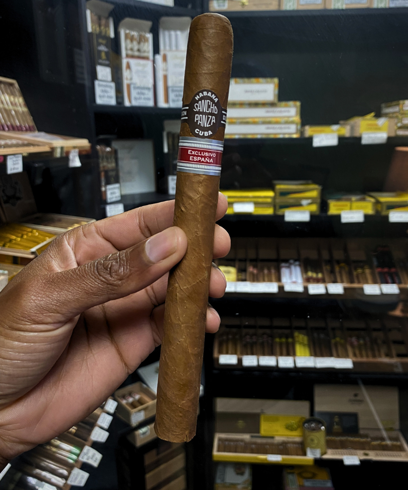 Cigar held by a hand with a store shelf of cigars in the background. Sancho Panza Quijote Regional Edition Spain