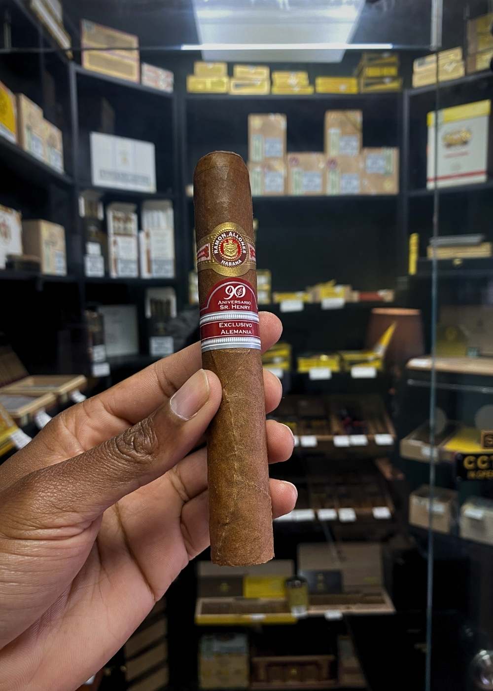 Cigar held by a hand with a store shelf of cigars in the background