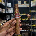 Cigar held by a hand with a store shelf of cigars in the background